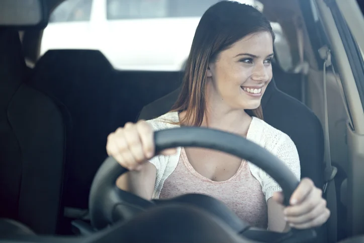 Com as mãos no volante, mulher sorri enquanto observa o trânsito. 