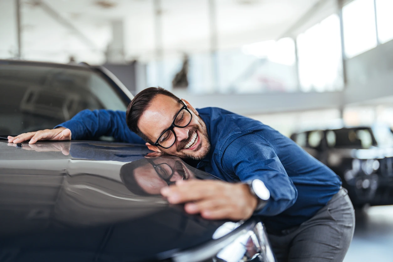 Carros confortáveis: descubra os melhores modelos para dirigir bem Homem feliz e sorridente em concessionária de carros, demonstrando satisfação ao comprar ou vender um veículo. Pessoa usando óculos de grau e camisa azul.