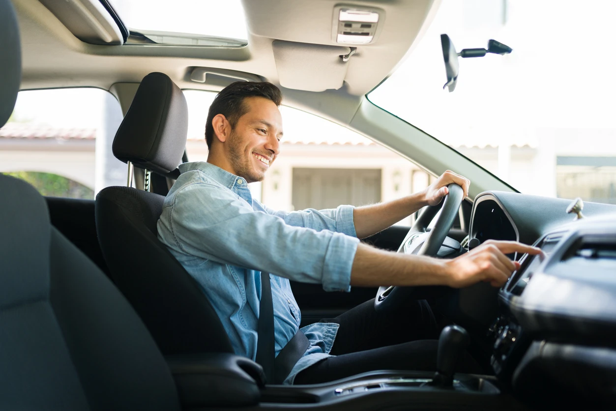 Homem sorridente dirigindo um carro na manhã, usando cinto de segurança, em ambiente de residência com céu azul ao fundo, representando segurança ao volante.