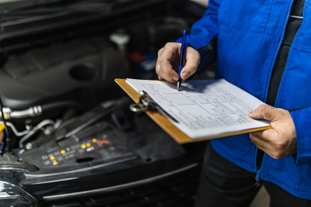 Profissional realizando inspeção técnica em veículo, verificando itens de segurança e condições do motor, em uma oficina de manutenção automotiva.