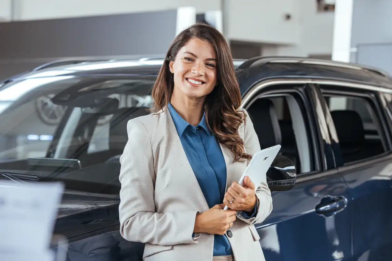 Mulher sorridente em uma concessionária de carros, segurando um smartphone, ao lado de um veículo novo, representando sucesso na compra de carros.