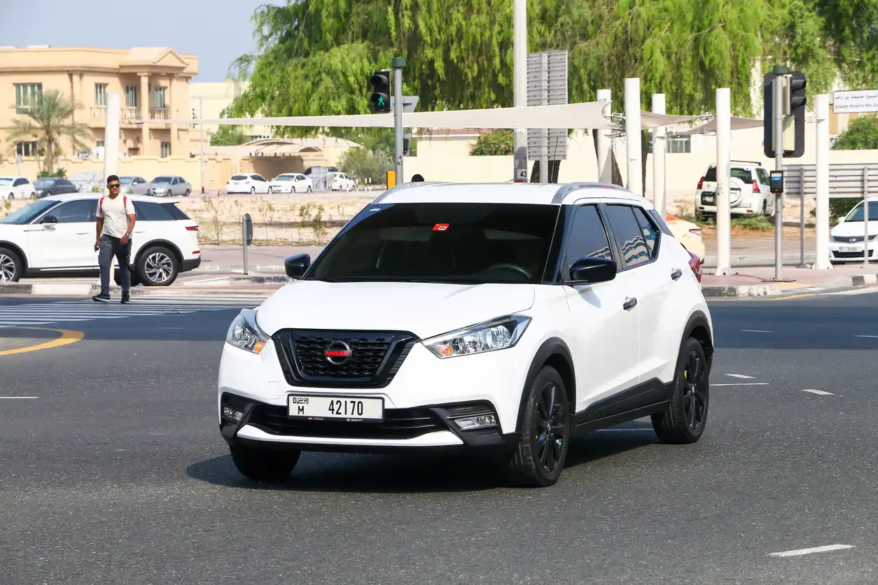 Nissan Kicks 2025: tudo que você precisa saber sobre o novo modelo Carro Nissan SUV branco circulando em uma rua movimentada durante o dia, com árvores e cruzamentos ao fundo, mostrando um veículo moderno e elegante.
